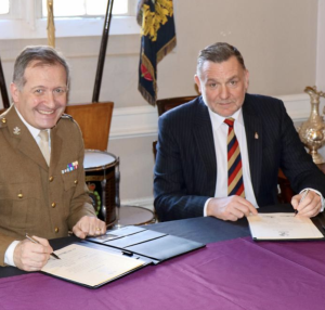 Two men signing papers at table- one in a suit the other in military garb. 